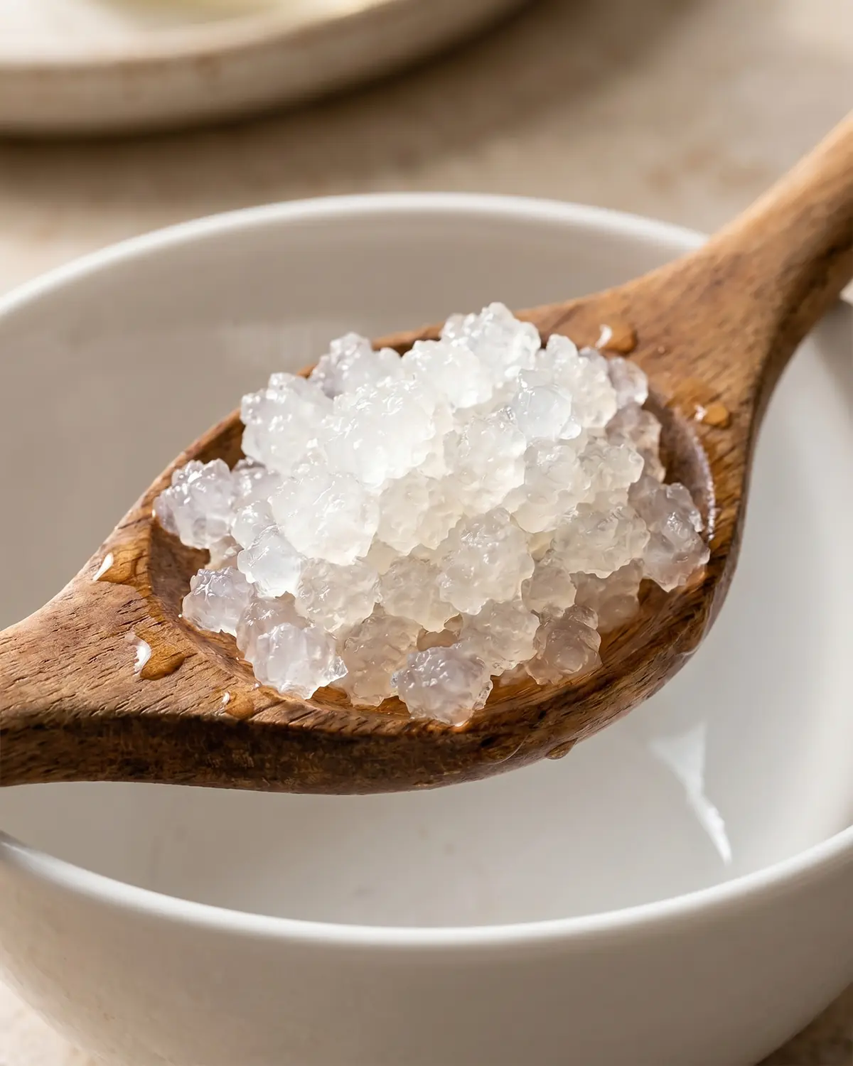Holzl&ouml;ffel mit durchscheinenden Wasserkefir-Kristallen &uuml;ber einer wei&szlig;en Schale in feuchter Nahaufnahme.