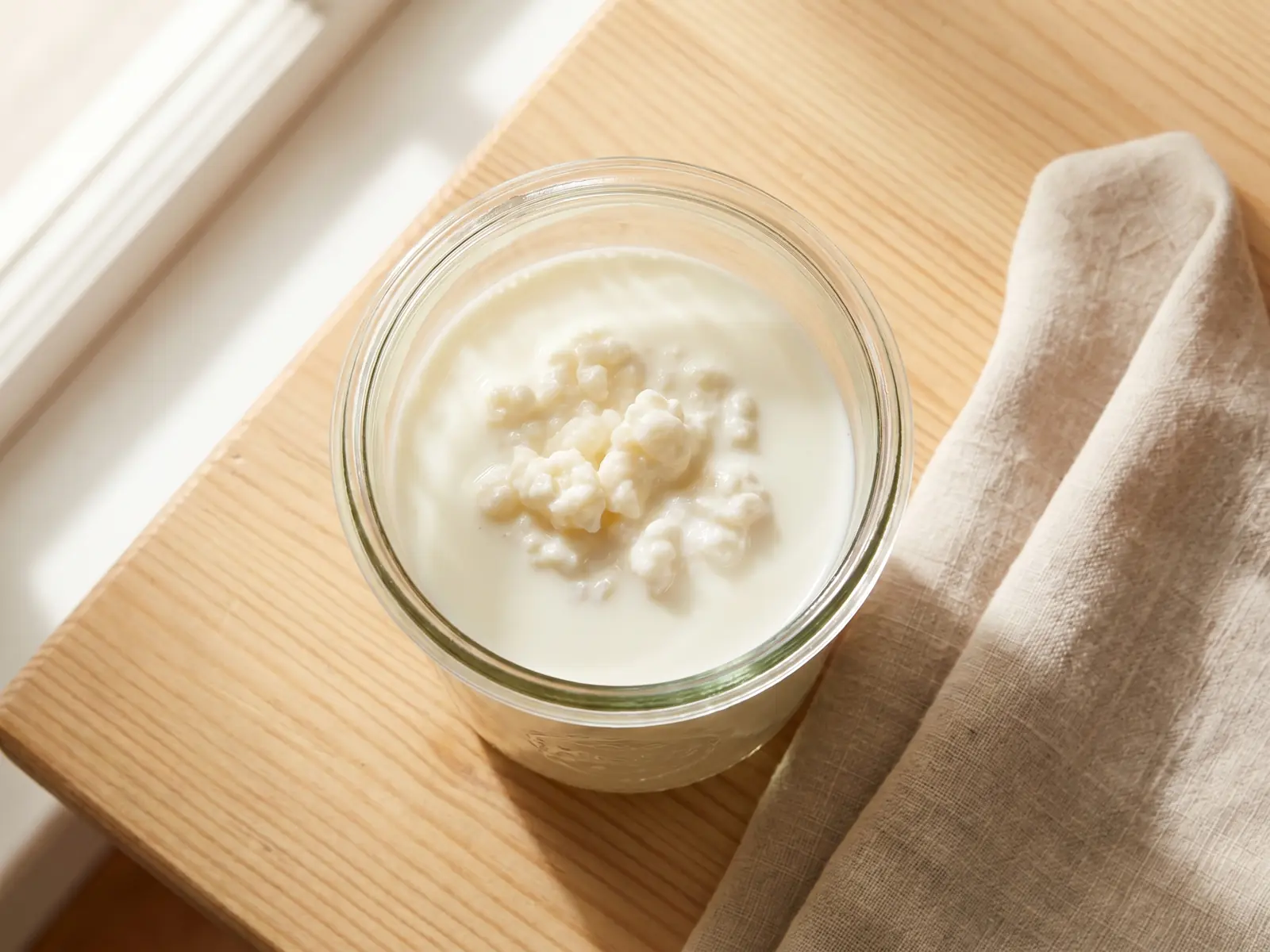 Glas mit fermentierendem Milchkefir neben Leinentuch auf einem Holztisch in ruhiger K&uuml;chenatmosph&auml;re