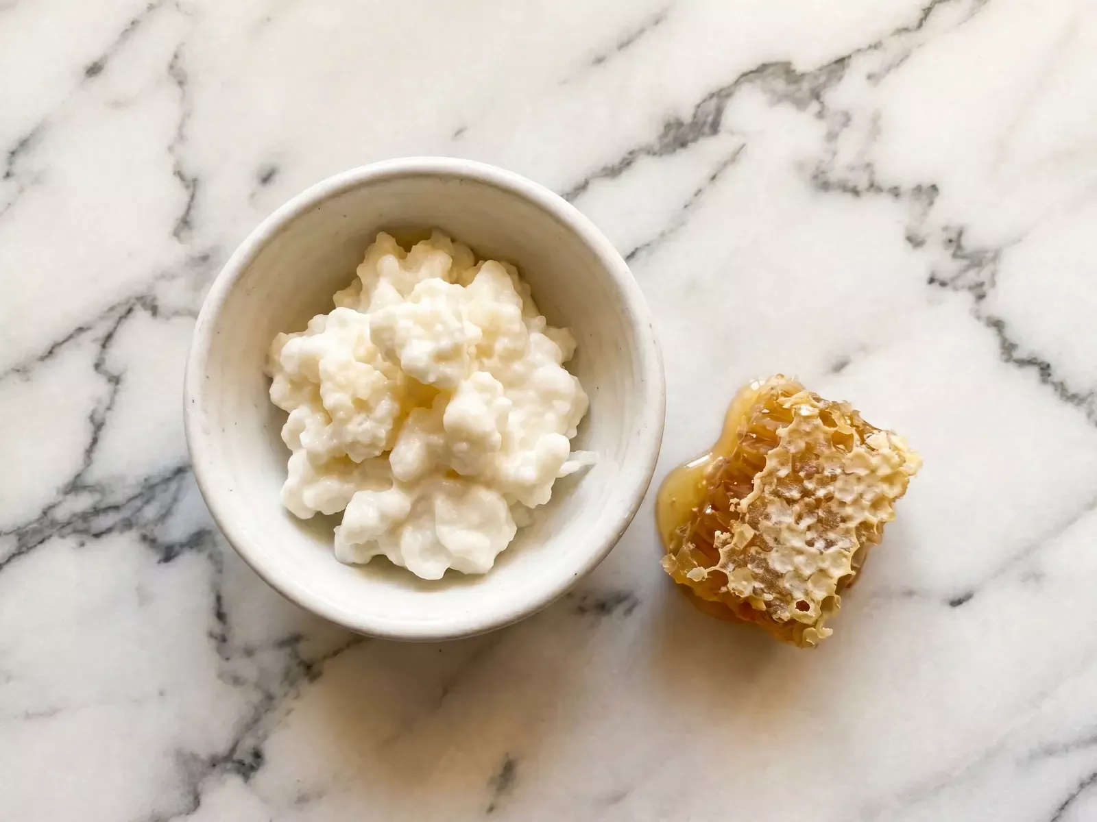 Schale mit nat&uuml;rlichem Milchkefir neben einer Honigwabe auf hellem Marmor in warmer Aufsicht.