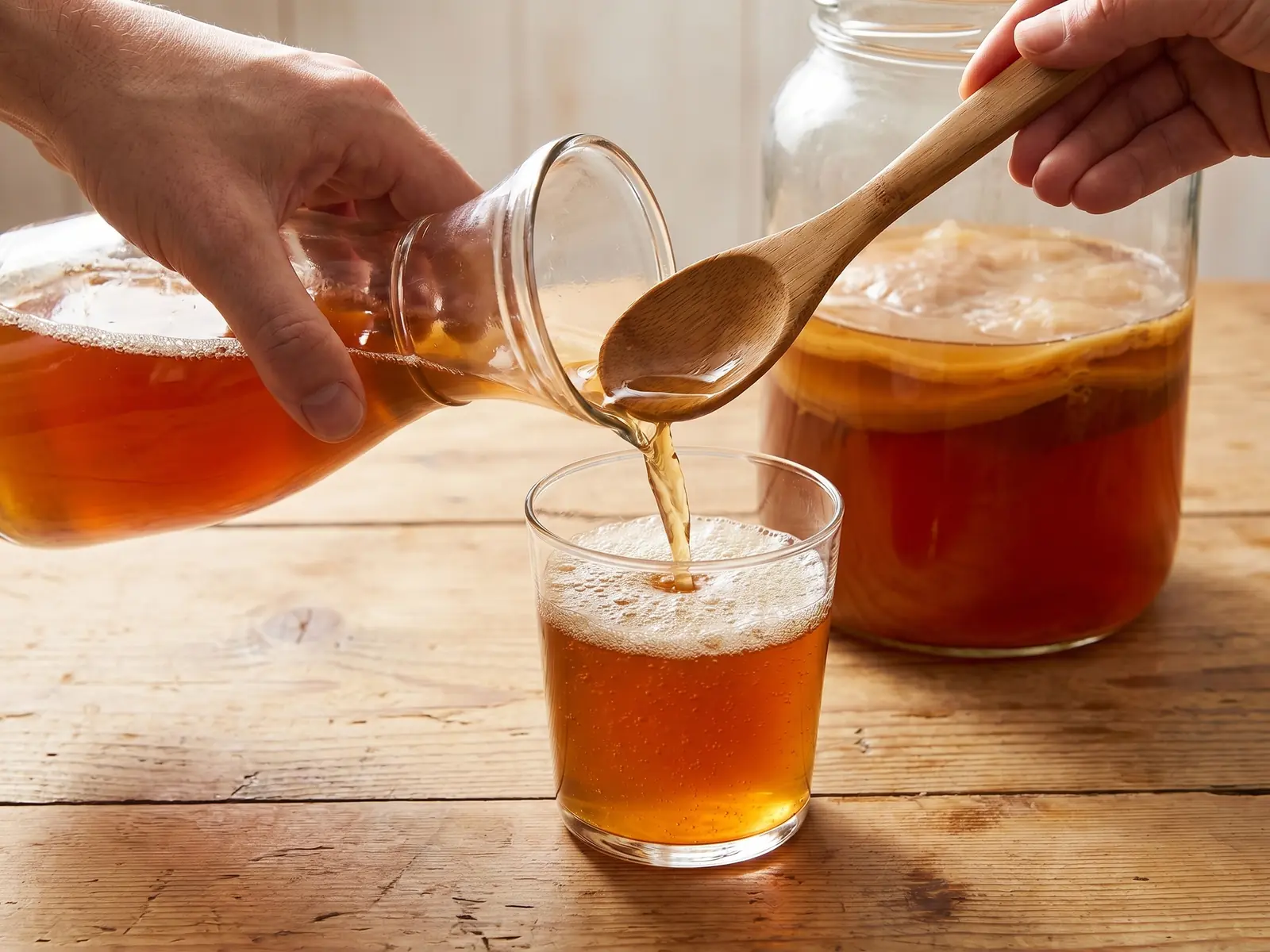 kombucha aufbewahren Eine Hand gießt fertigen Kombucha mit feinem Schaum in ein Glas auf einem hellen Holztisch.