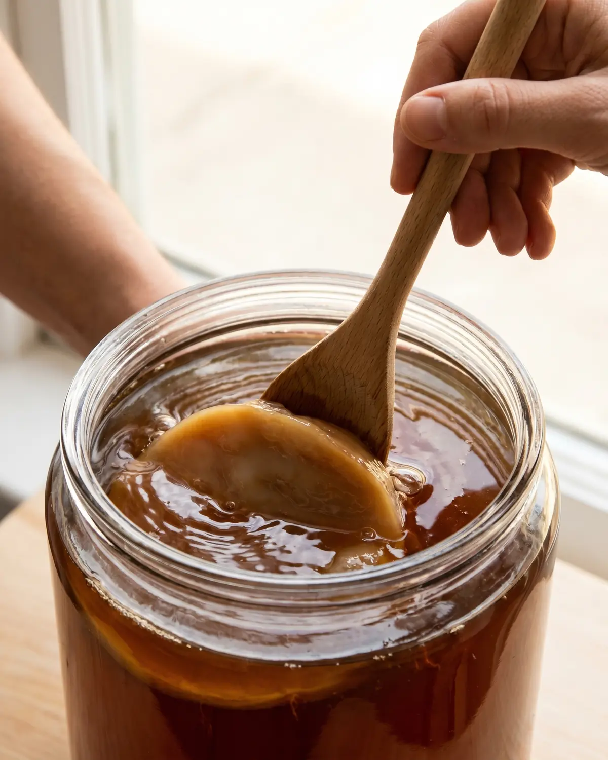 was ist kombucha Eine Holzlöffelspitze drückt den SCOBY behutsam unter bernsteinfarbenen Tee im Fermentationsgefäß.
