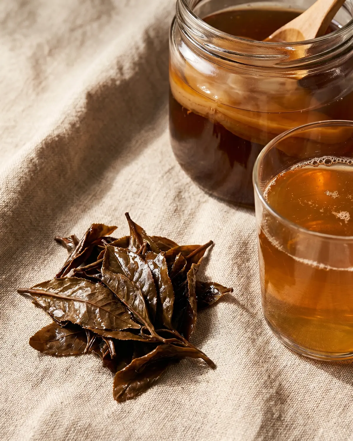 kombucha alkoholgehalt Feuchte Teeblätter liegen neben einem Glas Kombucha auf einer hellen Oberfläche aus rohem Leinen.