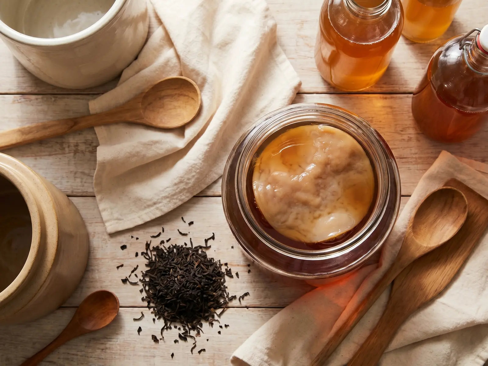 kombucha rezept fairment Cenitaler Kombucha-Ansatz mit SCOBY, Baumwolltuch, Teeblättern und hellem Holz in ruhiger Küchenatmosphäre.