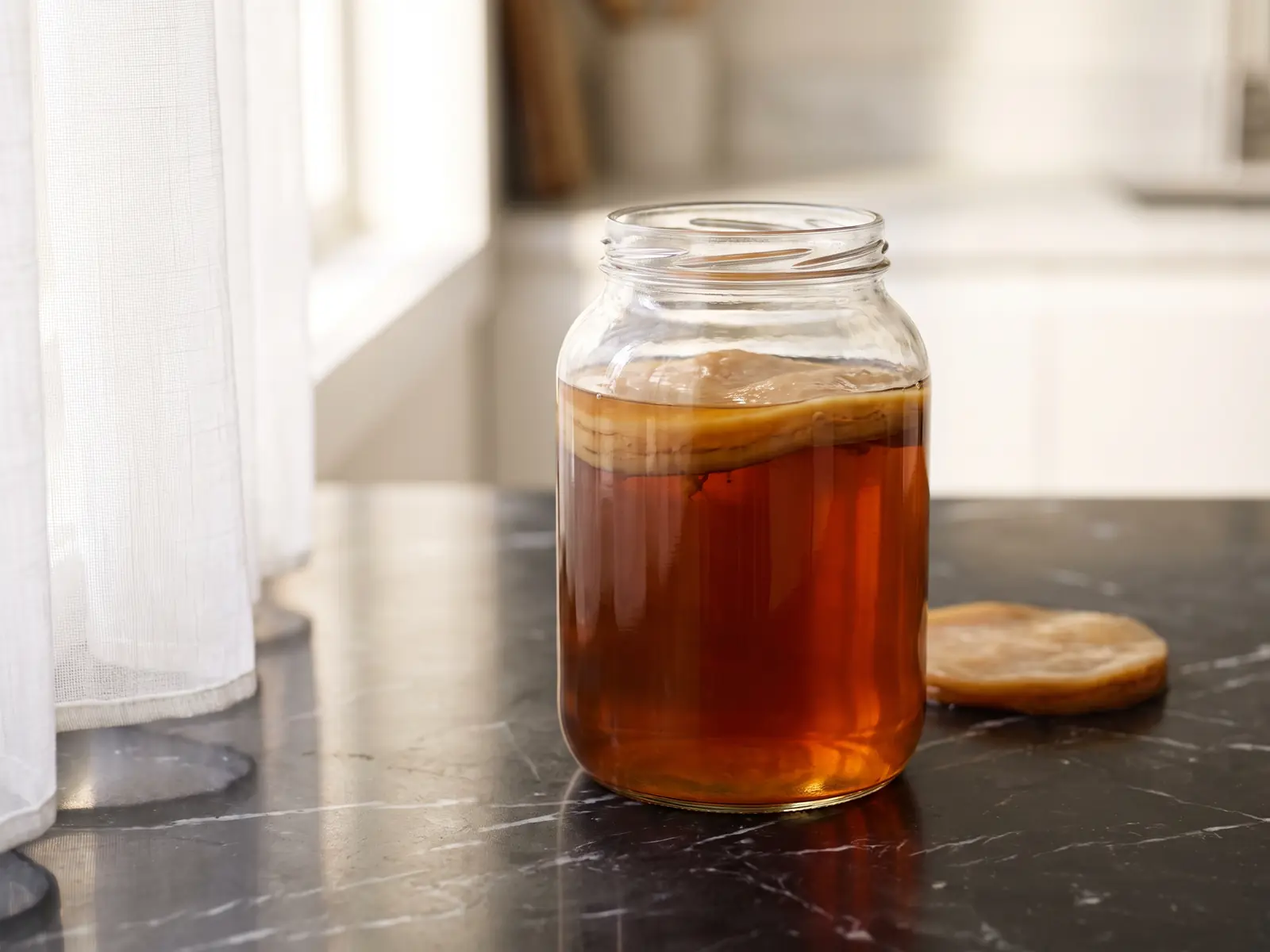 kombucha benefits Glas mit SCOBY und bernsteinfarbenem Tee auf schwarzem Marmor, im Hintergrund der Widerschein eines weißen Vorhangs.