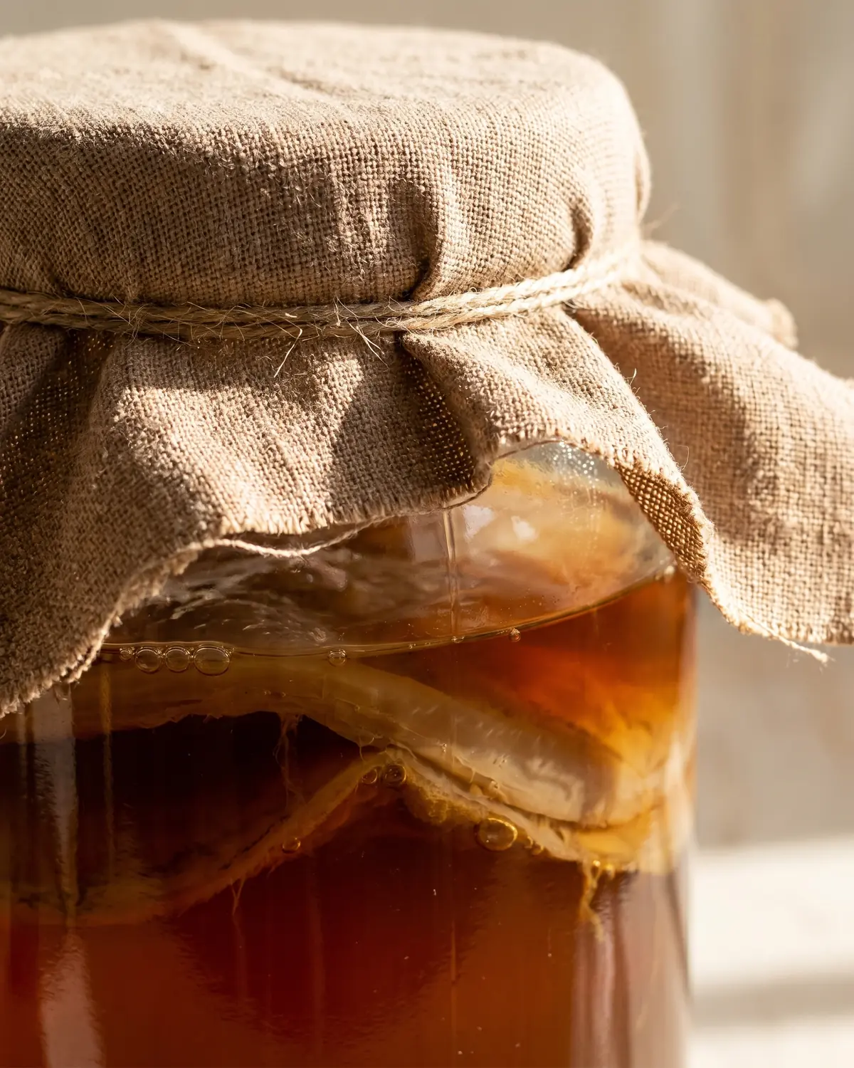 Nahaufnahme eines mit Baumwolltuch abgedeckten Kombucha-Glases, darunter ist dunkler Tee sichtbar.