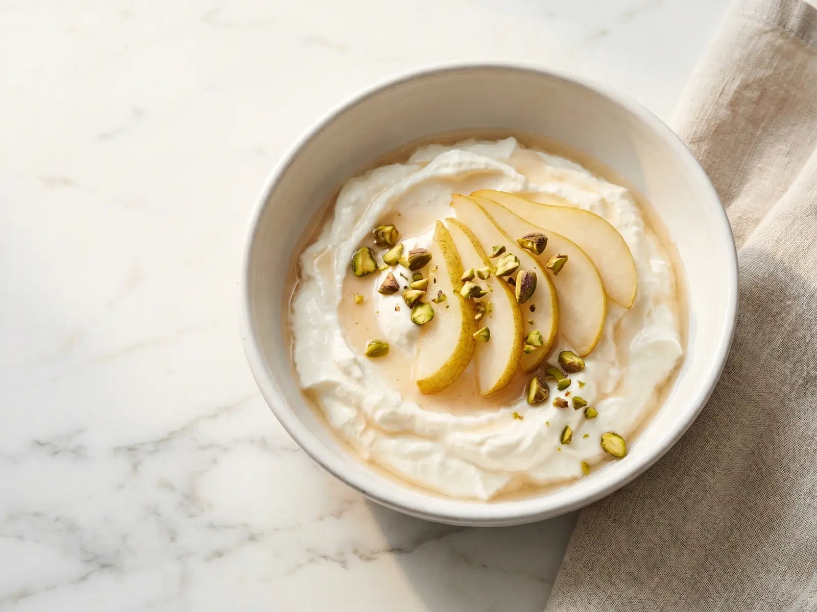 joghurt selber machen Cremiger Joghurt in einer weißen Keramikschale mit Birnenscheiben und Pistazien auf Marmor.