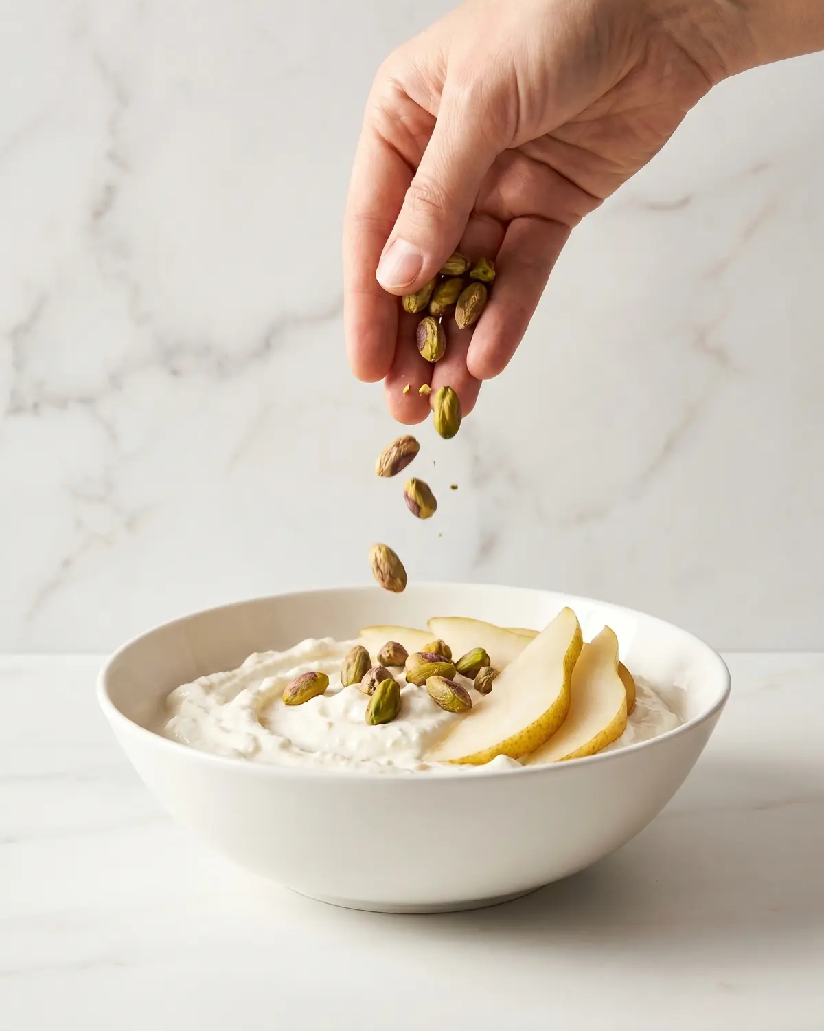 Eine Hand streut Pistazien &uuml;ber Joghurt mit Birne auf hellem Marmor, in ruhiger vertikaler Szene.